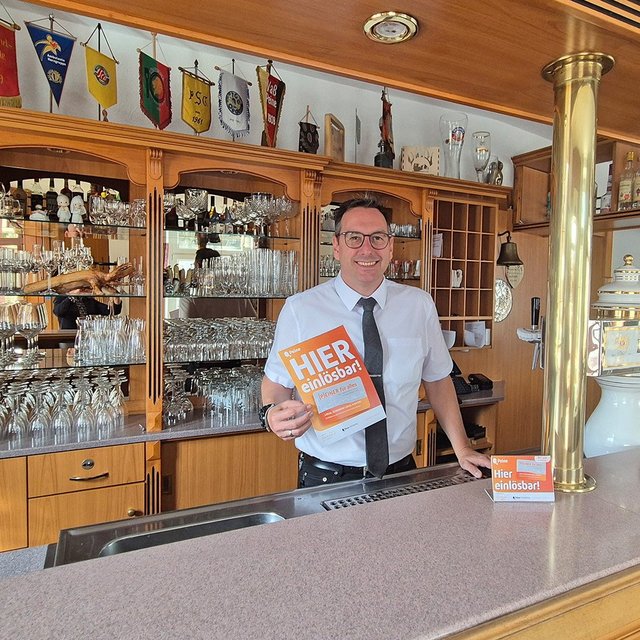 Ein Mann steht hinter einer Bar und hält ein orangefarbenes Schild mit der Aufschrift „HIER kostenlos“.