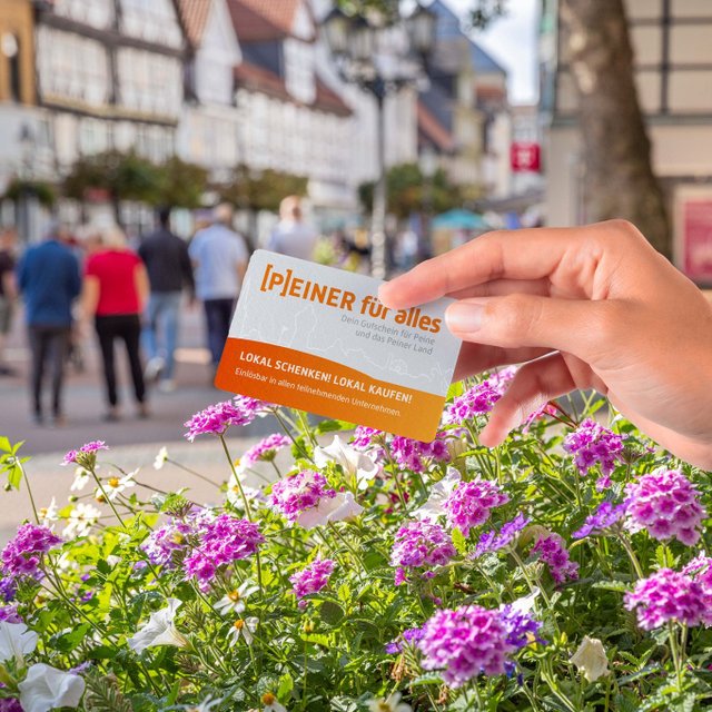 Eine Hand hält den Stadtgutschein „PEINER für alles“ über einem blühenden Blumenbeet in der Peiner Innenstadt. Im Hintergrund sind Fachwerkhäuser, Passanten und die Fußgängerzone unscharf zu sehen. Der Gutschein wirbt für lokales Schenken und Einkaufen in teilnehmenden Unternehmen in Peine.