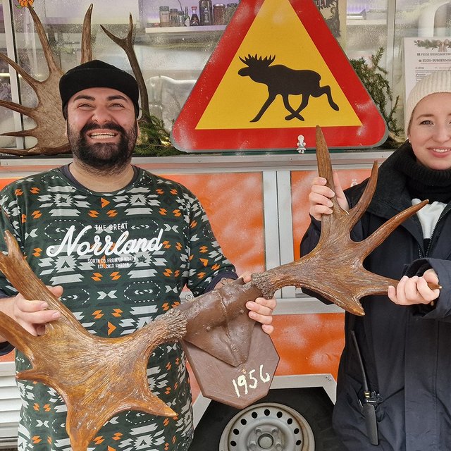 Ein Mann und eine Frau halten gemeinsam ein großes Geweih mit der Jahreszahl „1956“, im Hintergrund ein Warnschild mit Elch-Symbol an einem Foodtruck.