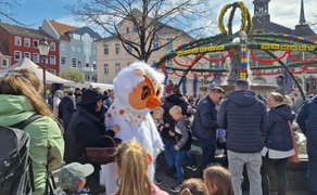 Viele Menschen versammeln sich auf dem Peiner Marktplatz rund um den bunt geschmückten Osterbrunnen, während das Maskottchen Uhlinchen Kinder begrüßt.