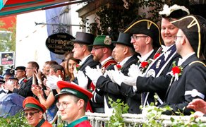 Mehrere Männer in historischen Uniformen mit Zylindern und weißen Handschuhen stehen an einem Geländer und klatschen.