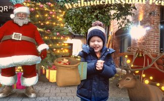 Augmented-Reality-Szene mit weihnachtlichem Motiv: In einer realen, städtischen Umgebung wird eine virtuelle Weihnachtswelt eingeblendet. Zu sehen sind eine digital dargestellte Figur des Weihnachtsmanns, ein geschmückter Weihnachtsbaum mit Lichtern und Geschenken sowie ein Rentier mit beleuchtetem Schlitten. Davor steht ein Kind in winterlicher Kleidung, das mit den virtuellen Elementen interagiert. Reale Architektur und Pflanzen bilden den Hintergrund und machen die Kombination aus digitaler Darstellung und realem Raum deutlich sichtbar.