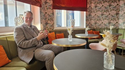 Mann sitzt in einem mit floraler Tapete, grüner Polsterbank und runden Tischen ausgestatteten Hotelzimmer mit orangefarbenen Kissen.