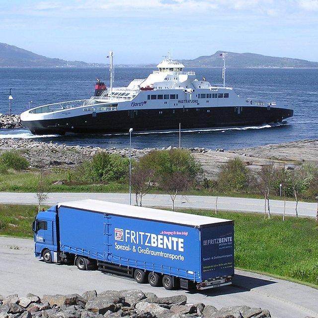 Lkw mit Aufschrift „FRITZ BENTE Spezial- & Großraumtransporte“ auf einer Straße am Wasser mit einer Fähre und Inseln im Hintergrund.