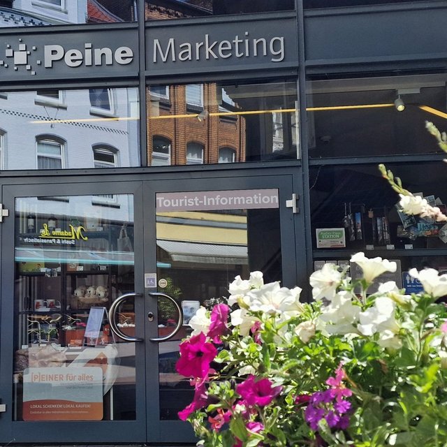 Eingang der Tourist-Information Peine im Gebäude „Peine Marketing“ mit Glasfassade und Beschilderung. Im Vordergrund stehen blühende Blumen, im Hintergrund sind Souvenirs und Informationsmaterial im Innenraum sichtbar.