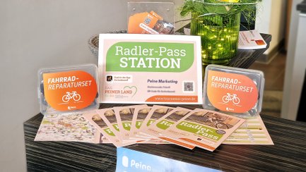 Zwei orangefarbene Fahrrad-Reparatursets neben Informationsmaterial und einem Schild mit der Aufschrift „Radler-Pass STATION“ auf einem Tisch.
