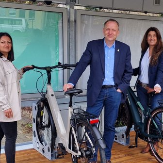Das Bild zeigt drei Menschen, die mit Fahrrädern an einem BikePort stehen. 