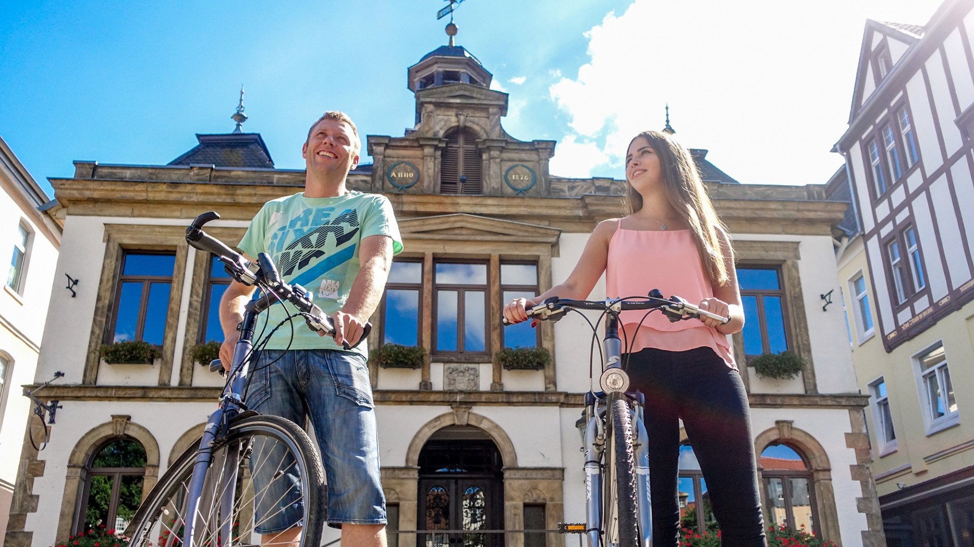 Zwei Personen mit Fahrrädern stehen vor einem historischen Gebäude mit großen Fenstern und einem Uhrenturm.