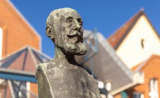 Nahaufnahme einer bronzenen Statue eines Mannes mit markantem Bart und ernstem Gesichtsausdruck. Die Figur ist detailreich gearbeitet und blickt leicht nach oben. Im Hintergrund sind unscharf Gebäude mit roten Ziegeldächern und ein klarer blauer Himmel zu sehen. Die Szene zeigt ein Denkmal im städtischen Raum bei sonnigem Wetter.