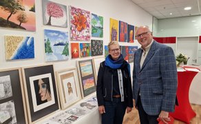 Eine Frau und ein Mann stehen vor einer Wand mit mehreren bunten Gemälden und Zeichnungen in einem Ausstellungsraum.