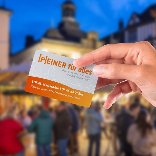 Hand hält eine Karte mit dem Text „Peiner für alles“ und „Lokal schenken, lokal kaufen!“ vor unscharfem Weihnachtsmarkt mit Menschen und Gebäuden.