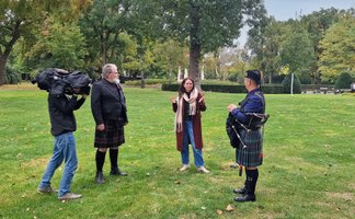 Ein Mann mit Kamera filmt zwei Männer im Kilt und eine Frau in einem Park.