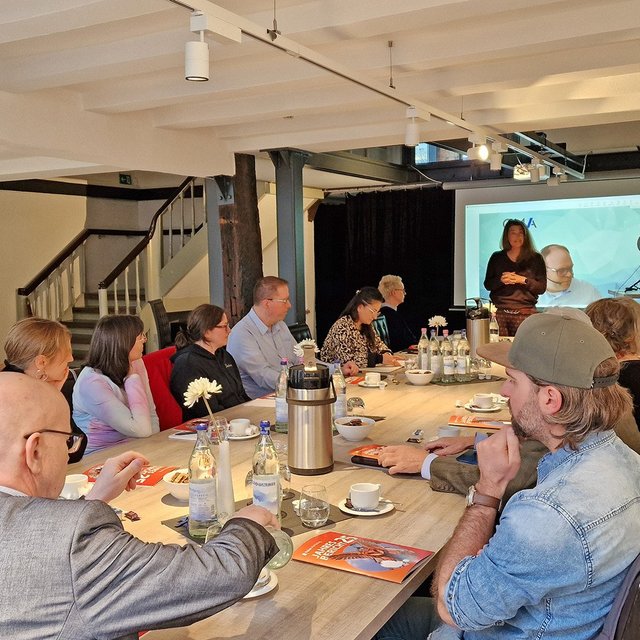 Menschen sitzen an einem langen Tisch im Peiner „Schwan“ mit sichtbaren Stahlträgern und hören Peine-Marketing-Geschäftsführerin Anja Barlen-Herbig zu, die vor einer Leinwand spricht.