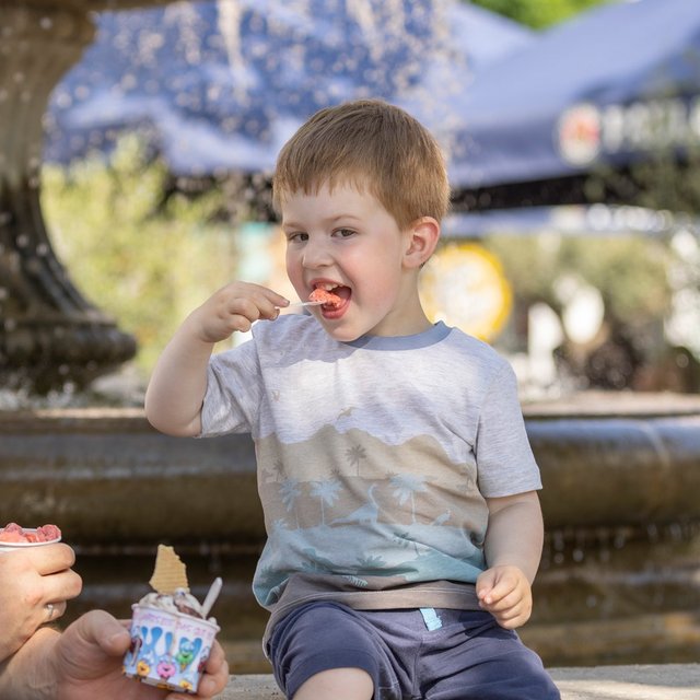 Kleines Kind isst Eis auf dem Marktplatz in Peine, während ein Erwachsener daneben sitzt – im Hintergrund ein Brunnen.