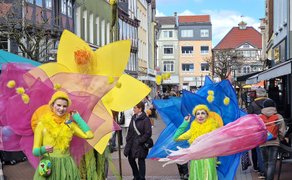 Zwei Personen in bunten Blumen-Kostümen mit großen Blütenblättern auf einer belebten Fußgängerstraße.