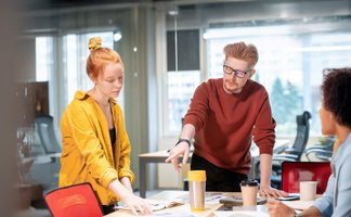 Drei junge Personen stehen um einen Tisch mit Papieren und Kaffeebechern in einem modernen Büro und diskutieren.