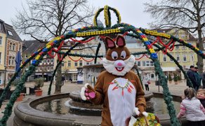 Eine als Osterhase verkleidete Person steht vor einem geschmückten Brunnen auf einem Marktplatz. Über dem Brunnen spannt sich eine grüne Girlandenkonstruktion, dekoriert mit bunten Eiern und Kugeln. Der Hase hält einen Korb mit Ostereiern und trägt eine Kette aus kleinen Möhren. Im Hintergrund sind historische Gebäude, einige Passanten sowie spielende Kinder zu sehen.