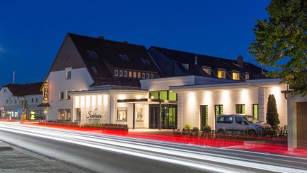 Außenansicht eines Hotels mit beleuchtetem Eingangsbereich und vorbeifahrendem Verkehr bei Nacht.
