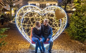 Paar sitzt auf roter Bank vor großem, leuchtendem Herz aus Lichterketten, umgeben von Tannenbäumen und Weihnachtsbeleuchtung.