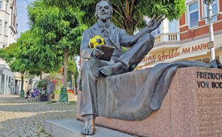 Das Bild zeigt die Skulptur von Friedrich von Bodenstedt auf dem Historischen Marktplatz, der einen gelben Fußbal in der Hand trägt.