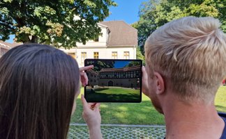 Zwei Personen betrachten auf einem Tablet eine digitale Rekonstruktion eines historischen Gebäudes im Park vor einem heutigen Bauwerk.