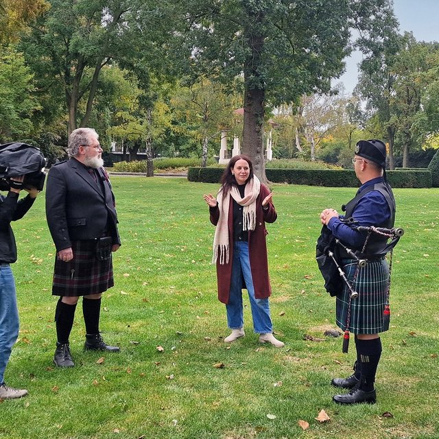 Ein Mann mit Kamera filmt zwei Männer im Kilt und eine Frau in einem Park.