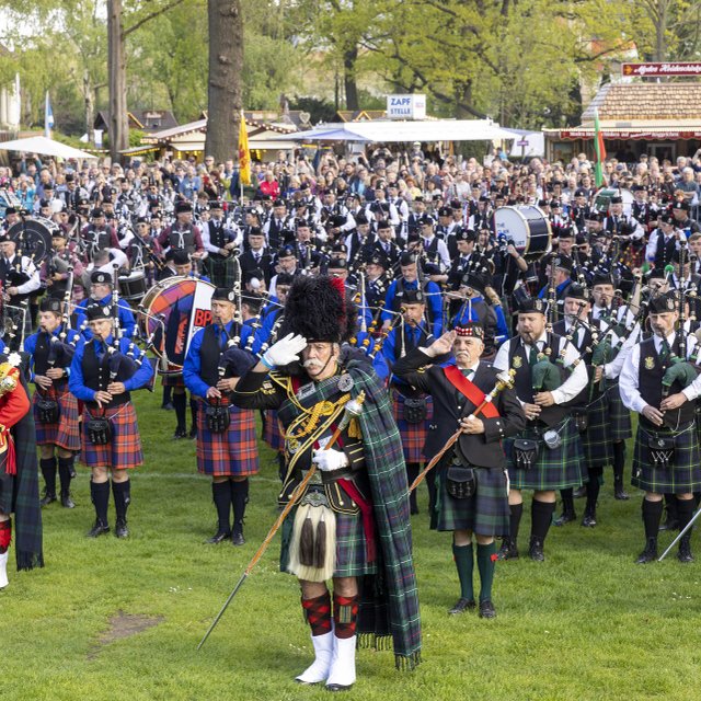 Großes Highland Gathering mit hunderten Dudelsackspielern in traditionellen Kilts, die gemeinsam auf einer Wiese musizieren, umgeben von zahlreichen Zuschauerinnen und Zuschauern bei einem schottischen Kulturfest.