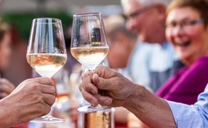 Anstoßen mit zwei Gläsern Weißwein beim Peiner Weinfest, Nahaufnahme von Händen mit Weingläsern vor unscharfem Hintergrund fröhlicher Gäste.