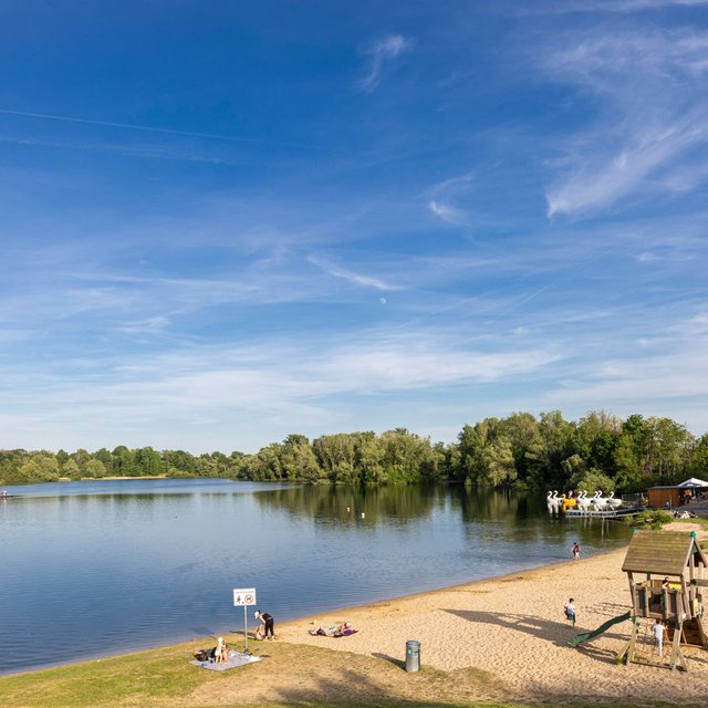 Badesee mit Sandstrand und Spielplatz an einem sonnigen Tag, Menschen entspannen am Ufer, Kinder spielen im Sand, im Hintergrund Bäume und ein Gebäude am Seeufer.