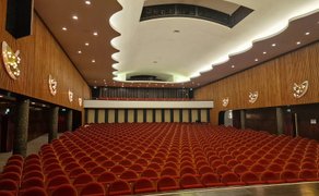 Leerer Theatersaal mit roten Sitzen, Holzverkleidung an den Wänden und geschwungener Decke mit Beleuchtung.