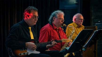 Drei Musiker des Ensembles Jazz53 spielen gemeinsam auf einer Bühne: Ein Gitarrist sitzt links mit E-Gitarre, in der Mitte spielt ein Saxofonist, rechts begleitet ein Pianist am Keyboard. Die Szene ist in warmes Bühnenlicht getaucht und vermittelt eine klassische Jazzclub-Atmosphäre.