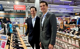 Zwei Männer stehen lächelnd nebeneinander in einem Elektronikmarkt. Im Vordergrund und Hintergrund sind verschiedene ausgestellte Elektrogeräte zu sehen.