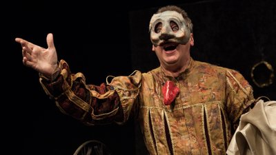 Ein Schauspieler in einem historischen Kostüm mit ausgestrecktem Arm auf einer dunklen Bühne.