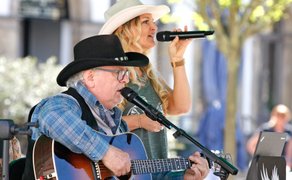 Zwei Musiker stehen auf einer Bühne im Freien und treten gemeinsam auf: ein Mann spielt Gitarre und singt ins Mikrofon, während eine Frau neben ihm mit Mikrofon in der Hand singt, beide im Country-Stil gekleidet.