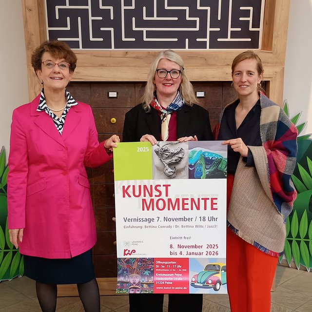 Drei Frauen stehen vor einer Holzwand mit Labyrinth-Optik und halten ein Plakat mit der Aufschrift „KUNST MOMENTE“ und „KiP“ in den Händen.