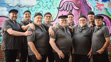 Mehrere Mitglieder des Männerensembles Kreischsägen 2.0 stehen eng beieinander und posieren lachend für ein Gruppenfoto. Die Sänger tragen einheitliche dunkle Poloshirts, Halstücher und schwarze Mützen. Im Hintergrund befindet sich eine bunt bemalte Graffitiwand.