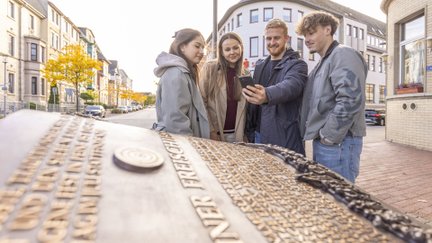 Vier junge Erwachsene entdecken Peine bei einer digitalen Adventuretour und schauen gemeinsam auf ein Smartphone an einer historischen Station in der Innenstadt.