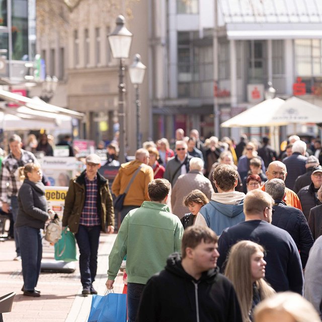Belebte Fußgängerzone in einer Innenstadt mit zahlreichen Menschen beim Einkaufen und Flanieren, Außengastronomie, Geschäften und Sonnenschirmen bei sonnigem Wetter.