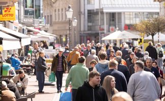 Belebte Fußgängerzone in einer Innenstadt mit zahlreichen Menschen beim Einkaufen und Flanieren, Außengastronomie, Geschäften und Sonnenschirmen bei sonnigem Wetter.
