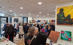 Menschenmenge in einer Galerie mit zahlreichen farbigen Gemälden an den Wänden, darunter ein großes Bild einer sitzenden Figur in Grün auf gelbem Hintergrund.