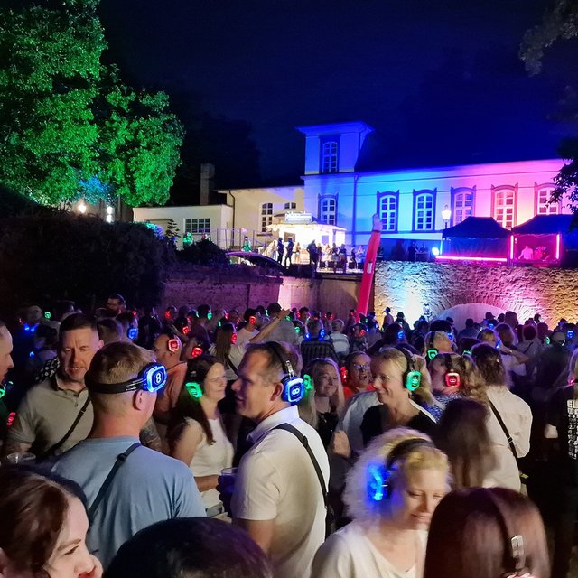 Große Menschenmenge mit leuchtenden Kopfhörern bei einer Open-Air-Party vor einem beleuchteten Gebäude in der Nacht.