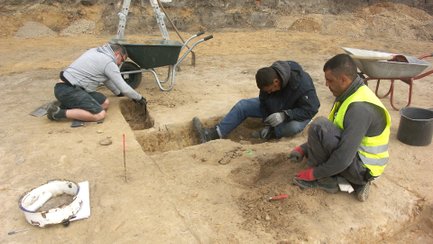 Drei Personen graben in einer archäologischen Ausgrabungsstätte in einer rechteckigen Grube im Boden.