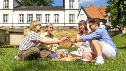 Vier Jugendliche sitzen auf einer Picknickdecke im Grünen, stoßen mit Getränken an und genießen gemeinsam ihre Ferien in Peine.