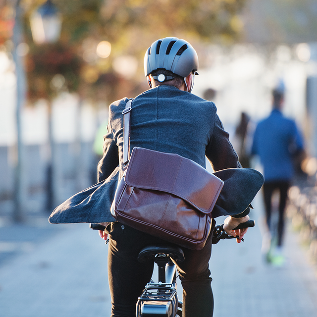 Person mit Fahrradhelm und Umhängetasche fährt auf einem Radweg entlang einer Baumreihe und Parkbänken.