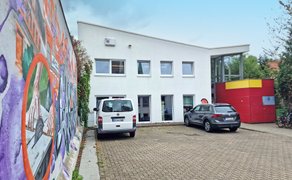 Gebäude mit weißer Fassade und rotem, gelbem Anbau, davor zwei geparkte Autos, links bunte Wand mit Graffiti.