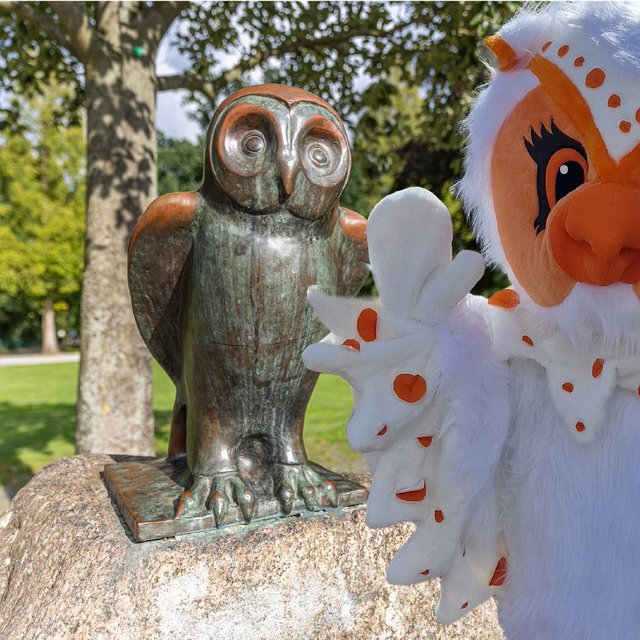 Uhlinchen steht neben einer Eulenstatue im Grünen in Peine und winkt freundlich in die Kamera.