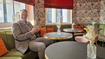 Mann sitzt in einem mit floraler Tapete, grüner Polsterbank und runden Tischen ausgestatteten Hotelzimmer mit orangefarbenen Kissen.