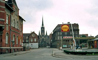 Eine Straße mit alten Backsteinhäusern und einer Shell-Tankstelle, im Hintergrund eine Kirche.