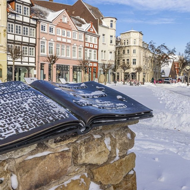 Verschneiter Marktplatz in Peine mit historischer Häuserzeile und offenem Buchdenkmal im Vordergrund bei blauem Winterhimmel.