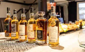 Whiskyflaschen unterschiedlicher Marken und Formen stehen in einer Reihe auf einem hellen Holztisch mit schwarzem Muster im Hintergrund eines Gastraums.